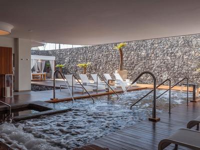 Indoor pool with jacuzzi and sun loungers in a modern hotel area with a stone wall.