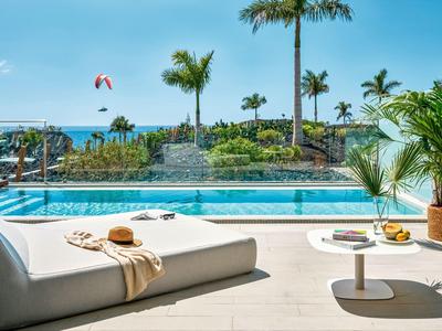 Moderní bazénová oblast s lehátkem, stolem a tropickou zahradou pod jasnou oblohou.