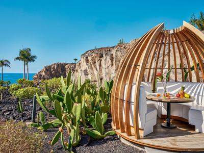 Wooden cabin with seating area surrounded by cacti, overlooking rocks and sea.