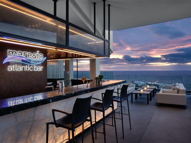 Moderní bar s pultem a sezením s výhledem na moře při západu slunce.