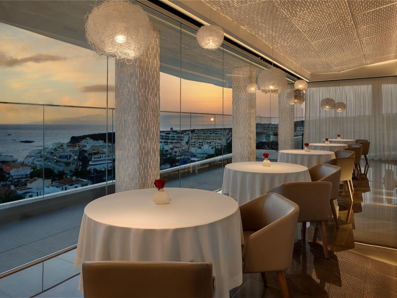 Elegantní restaurace s kulatými stoly a výhledem na město při západu slunce.