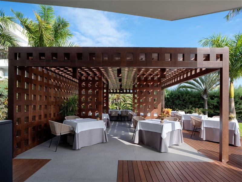 Elegantní restaurace s bílými prostřenými stoly pod dřevěnou pergolou a palmami v pozadí.