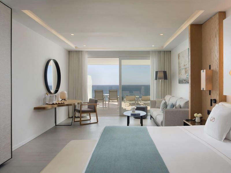 Moderně zařízený hotelový pokoj se manželskou postelí, psacím stolem, židlí a výhledem na moře.