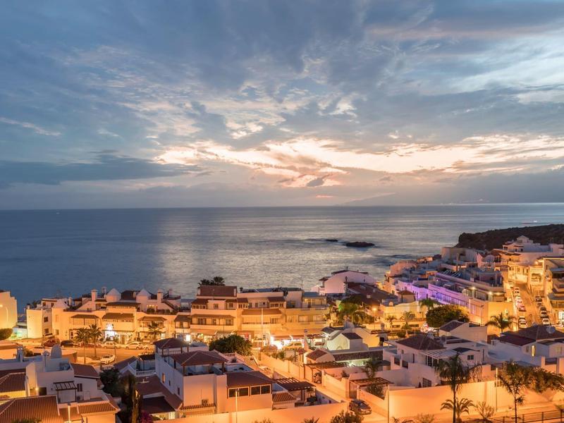 Pohled na pobřežní město s osvětlenými budovami a klidným mořem při západu slunce.