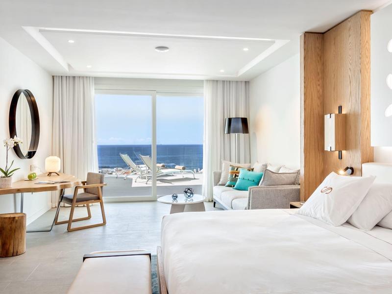Moderní hotelový pokoj s výhledem na moře, bílými zdmi, dřevěnými prvky a stylovým zařízením.