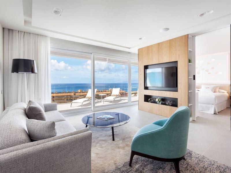 Modern hotel room with seating area and ocean view through large glass windows.