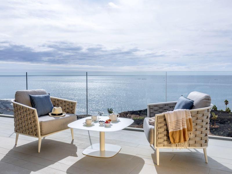 Two comfy chairs with cushions and throws on terrace overlooking sea under cloudy sky.