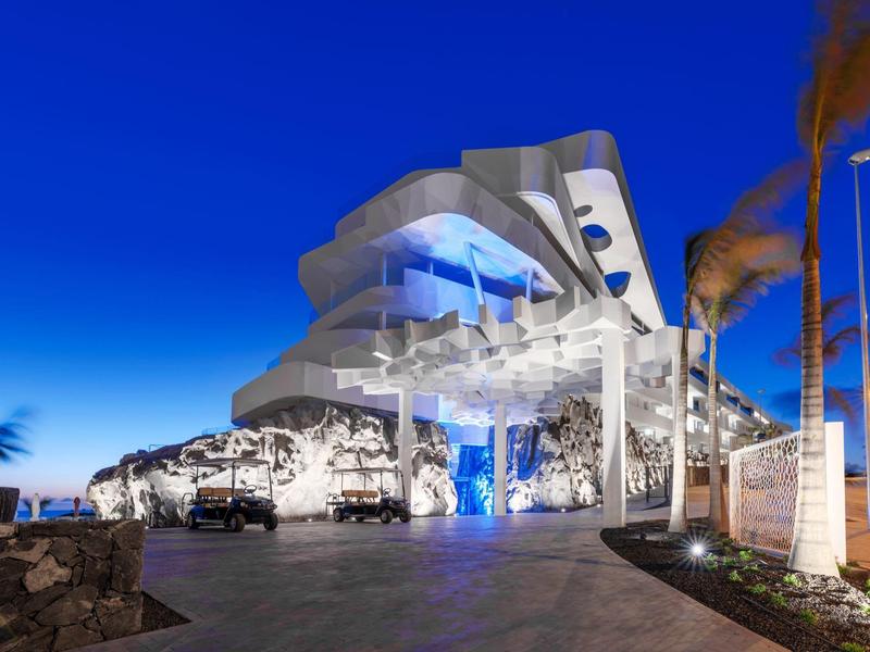 Modern hotel with illuminated terrace and palm trees at night under a clear sky.