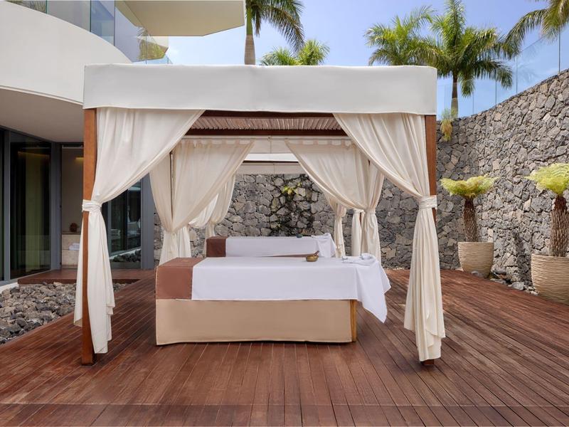 Elegant canopy bed on wooden deck, surrounded by stones and palm trees.