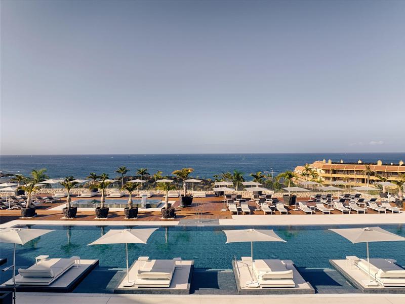 View of a pool with lounge chairs and the sea in the background on a sunny day.