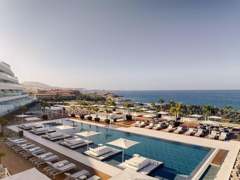 Modern hotel pool with sun loungers, sea view, and sunny sky.