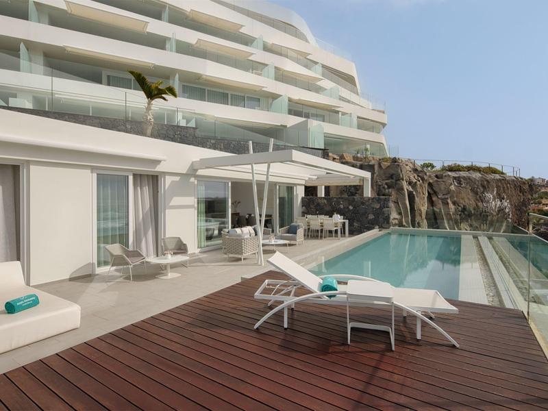 Modern hotel balcony with lounge chairs, umbrella, and pool overlooking the sea.