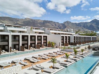 Nowoczesny kompleks hotelowy z wieloma basenami i leżakami na tle gór pod czystym niebem.