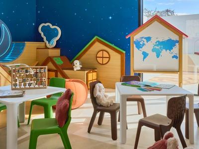 Strefa dla dzieci z kolorowymi krzesłami, stolikami i drewnianymi zabawkami przed niebieską ścianą z wzorami gwiazd.