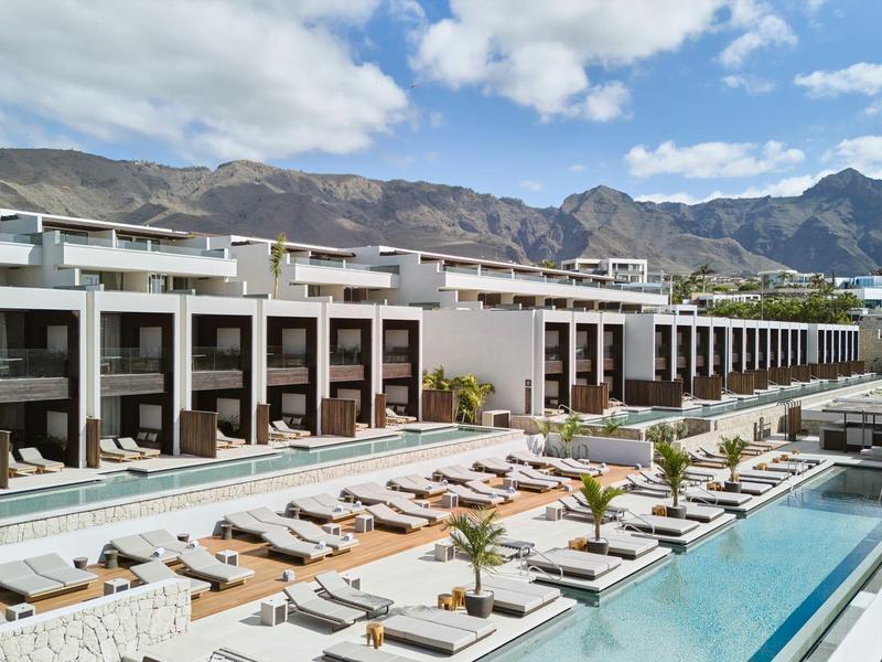 Nowoczesny kompleks hotelowy z wieloma basenami i leżakami na tle gór pod czystym niebem.