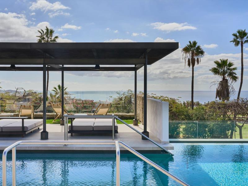 Nowoczesny basen zewnętrzny z widokiem na morze, meble lounge i palmy pod niebieskim niebem.