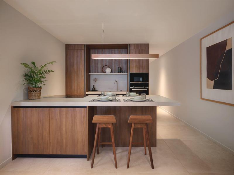 Nowoczesna kuchnia z drewnianymi frontami, wyspą i dwoma drewnianymi stołkami barowymi w jasnym pomieszczeniu.