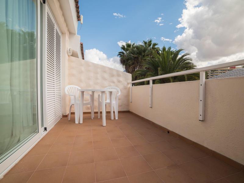 Balcone con pavimento marrone, pareti bianche, un tavolo bianco e tre sedie bianche.