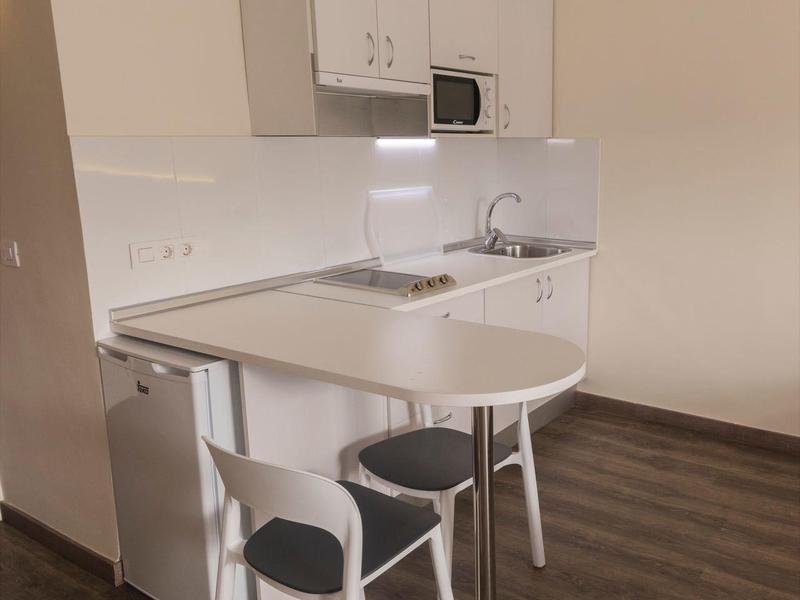 Piccola angolo cottura con tavolo, due sedie e apparecchiature da cucina minime in una stanza luminosa.