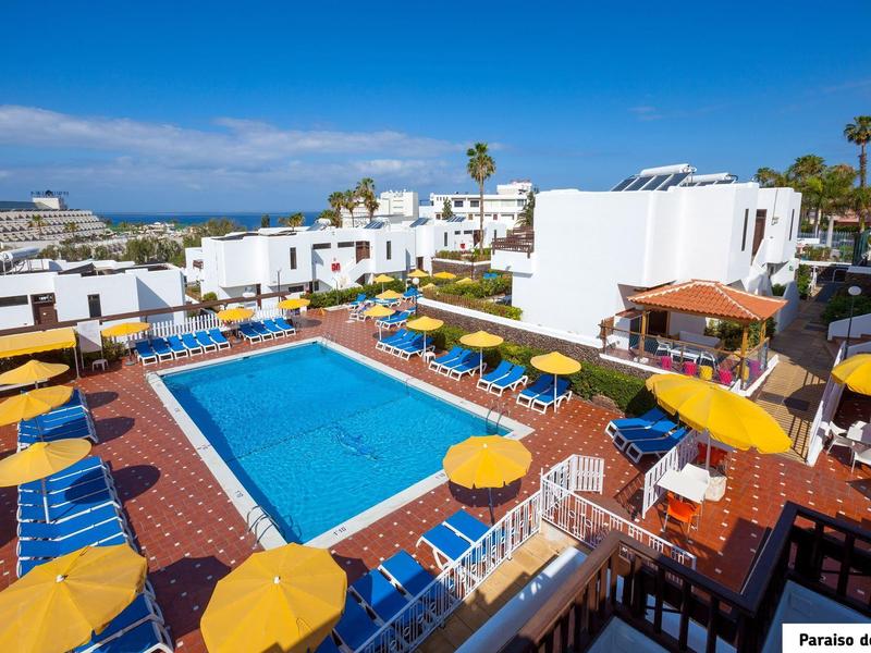 Area piscina dell'hotel con lettini blu e ombrelloni gialli circondata da architettura bianca.