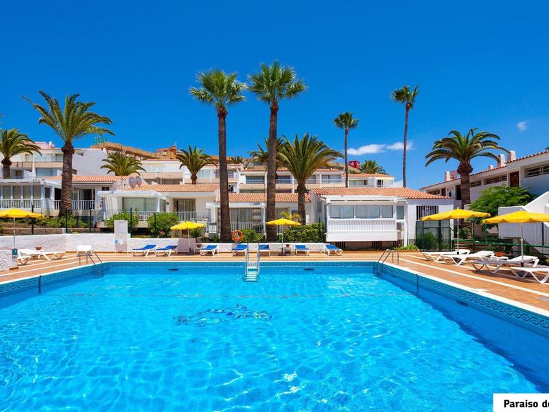 Grande piscina all'aperto davanti a case vacanza bianche e palme sotto un cielo azzurro limpido.