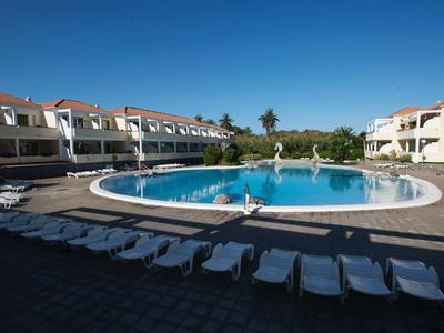 Round swimming pool with sun loungers in front of hotel buildings under clear blue sky.
