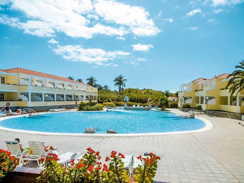 Large round pool with lounge chairs and flowers, surrounded by hotel buildings and palm trees.