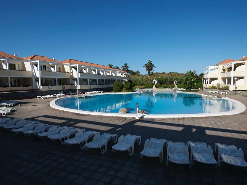Round swimming pool with sun loungers in front of hotel buildings under clear blue sky.