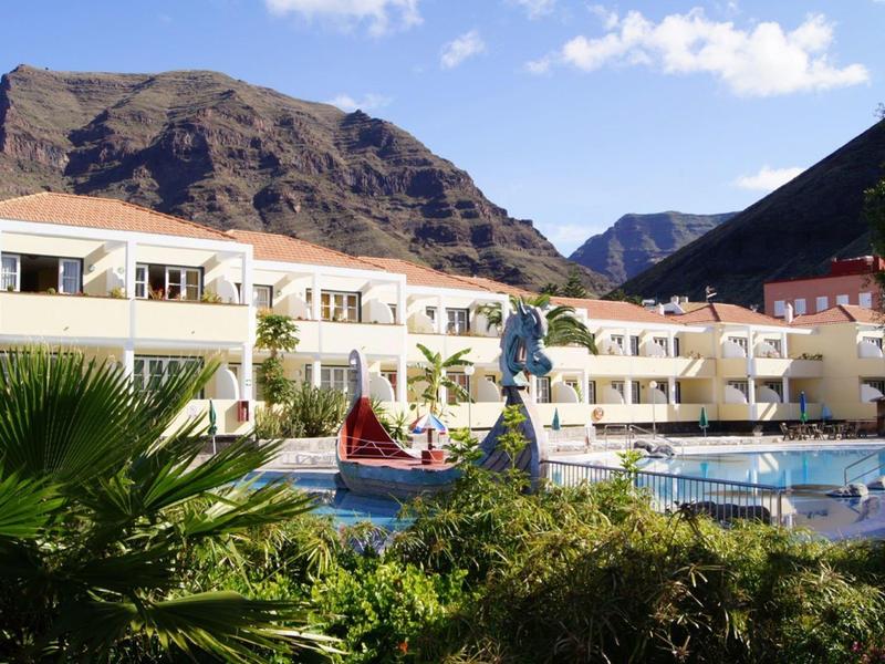 Hotel with pool in front of mountains and lush vegetation under sunny sky