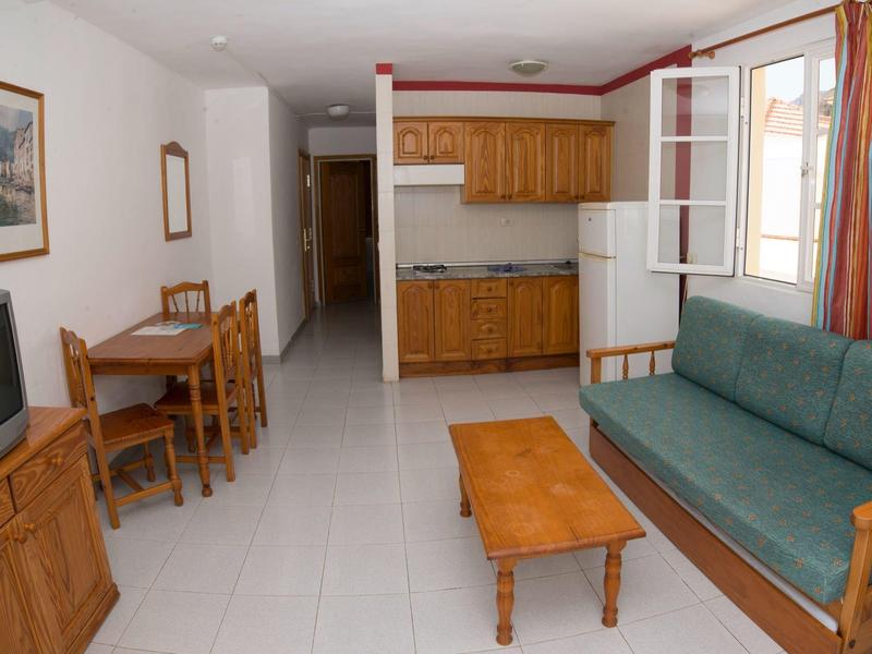 Compact living area with sofa, wooden table, dining set, TV, and kitchenette.
