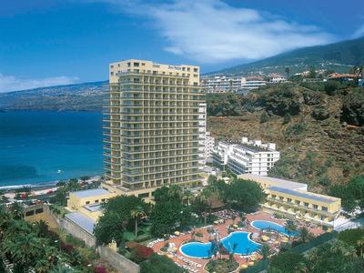 Hoher Hotelbau mit runden Pools, umgeben von Palmen an Küste mit blauem Meer und Bergen im Hintergrund.
