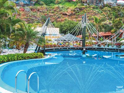 Blauer Pool mit Metallleiter, hängender Fußgängerbrücke und bewaldetem Hügel im Hintergrund.