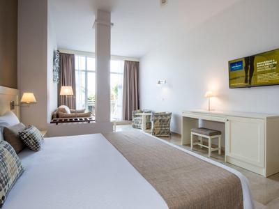Helles Schlafzimmer mit großem Bett, weißen Möbeln, zwei Sesseln und Fernseher an der Wand.