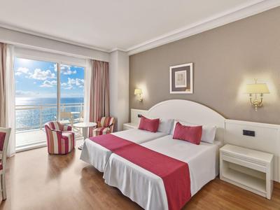 Een lichte hotelkamer met tweepersoonsbed, rode accenten en balkon met uitzicht op zee.