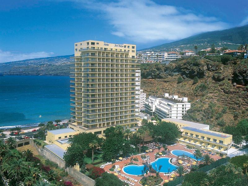 Hoog hotelcomplex met zwembaden, omringd door palmbomen, dicht bij de zee en bergen op de achtergrond.