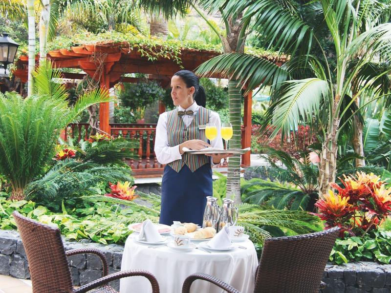 Serveerster serveert drankjes aan een buitentafel in een tropische hotel tuin.