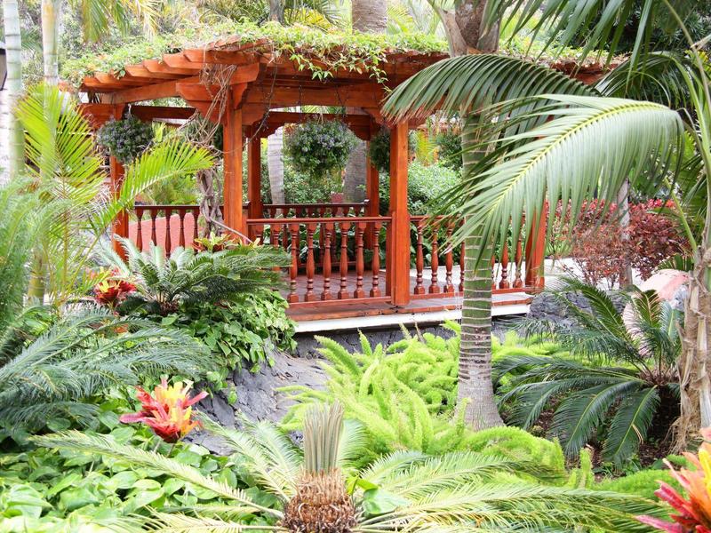 Een houten prieel omgeven door tropische planten en palmbomen in een tuin.