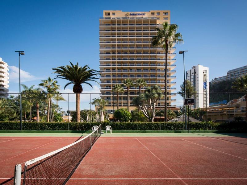Tennisbaan voor een hoog hotelgebouw met palmbomen en blauwe lucht.