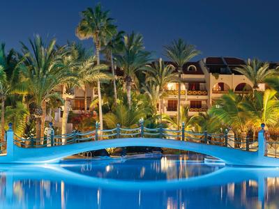 Illuminated bridge over a pool in front of a hotel at night with palm trees.