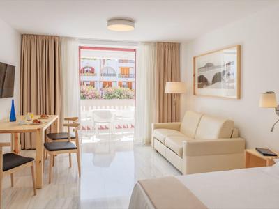 Camera d'albergo luminosa con balcone, arredi moderni e zona pranzo con tavolo e sedie.