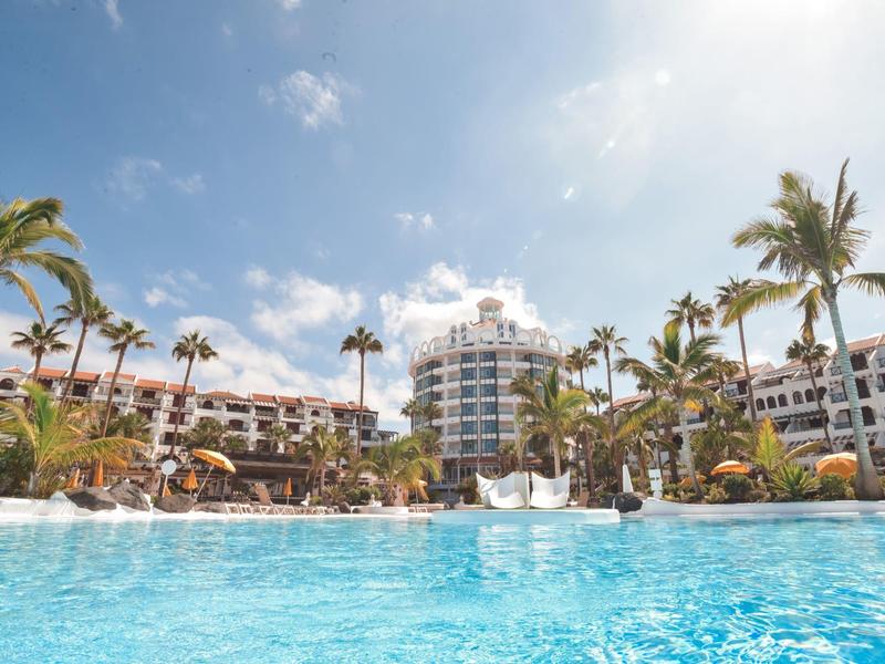 Grande piscina dell'hotel circondata da palme e aree esterne sotto un cielo soleggiato.