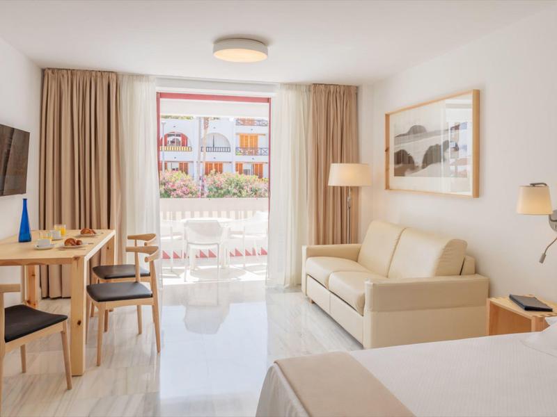 Camera d'albergo luminosa con balcone, arredi moderni e zona pranzo con tavolo e sedie.