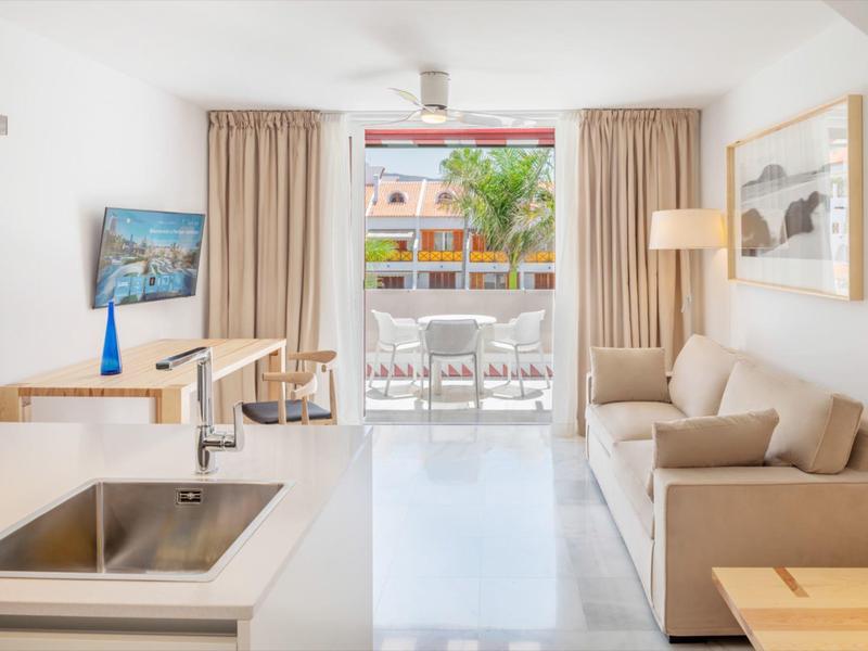 Spazio abitativo luminoso con divano, tavolo da pranzo e balcone con vista sulle palme.