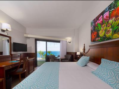 Bright hotel room with double bed, desk, TV, and balcony overlooking the sea.