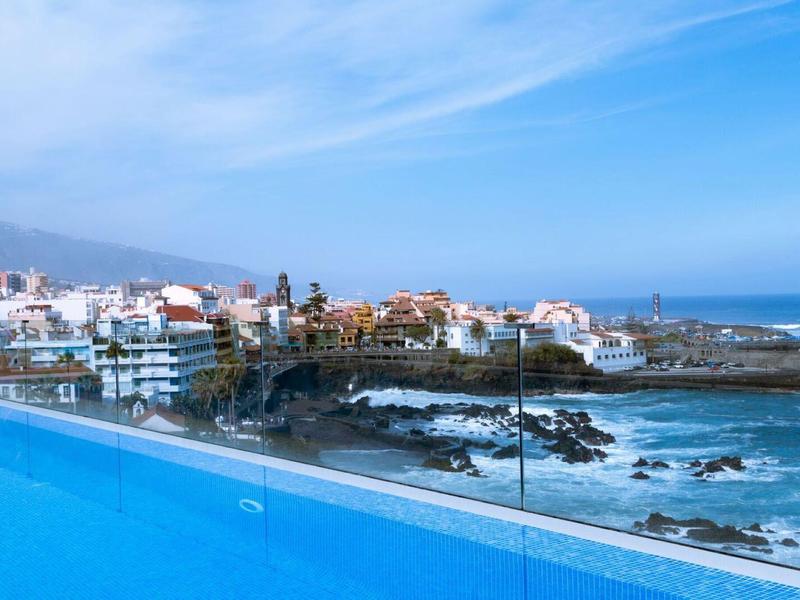 Blue pool overlooking coastal town and rocks in sea under cloudy sky.