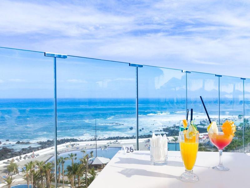 Two colorful cocktails on a white table with sea view behind glass railing.