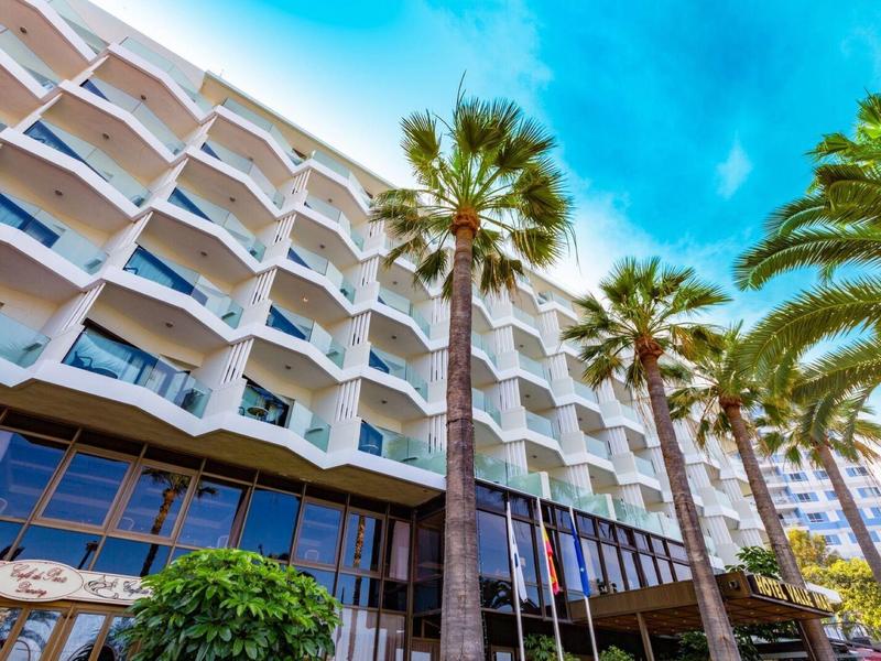 View of a modern hotel building with white balconies and palm trees in the foreground.