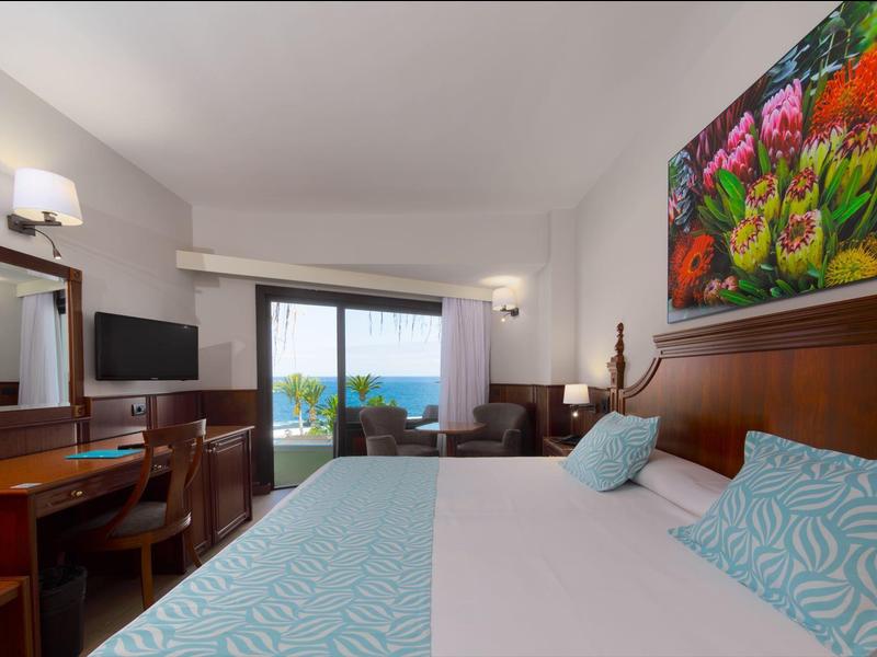 Bright hotel room with double bed, desk, TV, and balcony overlooking the sea.