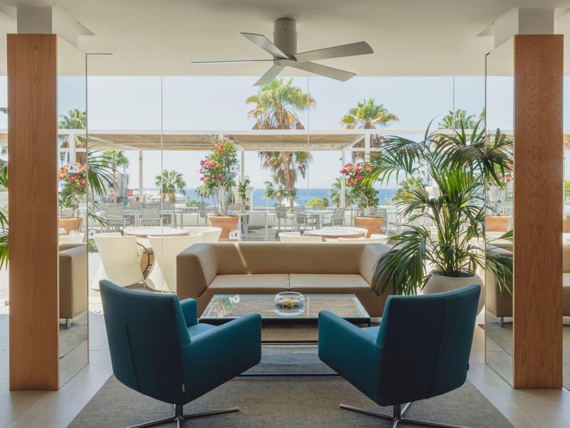 Modernes Wohnzimmer mit blauen Sesseln, Sofa, großen Fenstern und Palmenblick draußen.