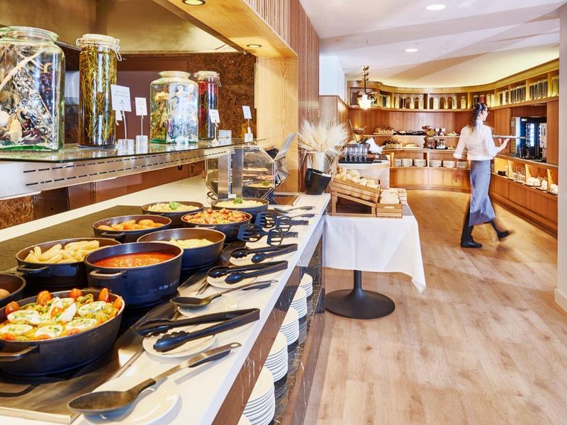 Buffet mit verschiedenen warmen Gerichten in schwarzen Schalen und Holzimitat-Boden in hellem Raum.